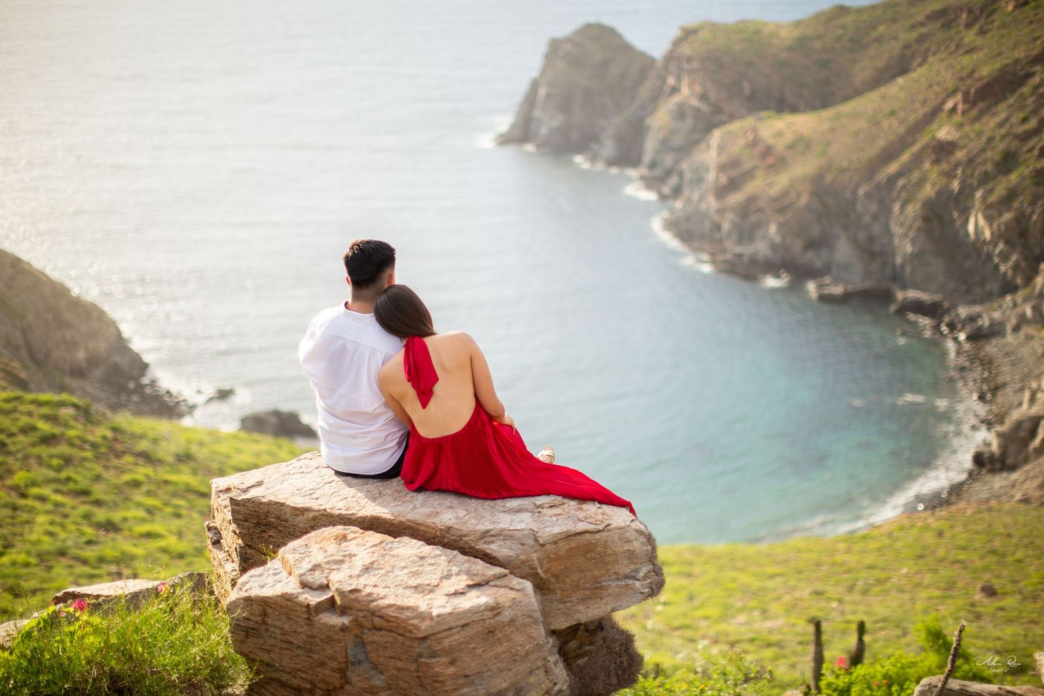 Couple sitting together on rocky cliff overlooking a serene blue bay.