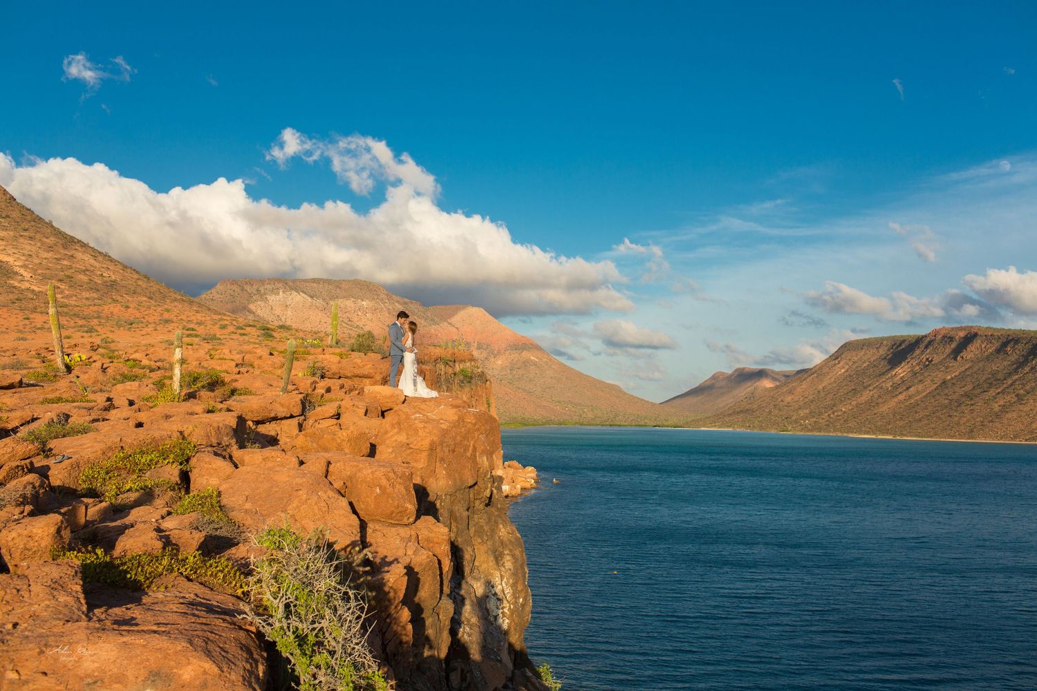 Bride and groom embrace on red rocky cliff overlooking turquoise sea with mountains and blue sky backdrop.