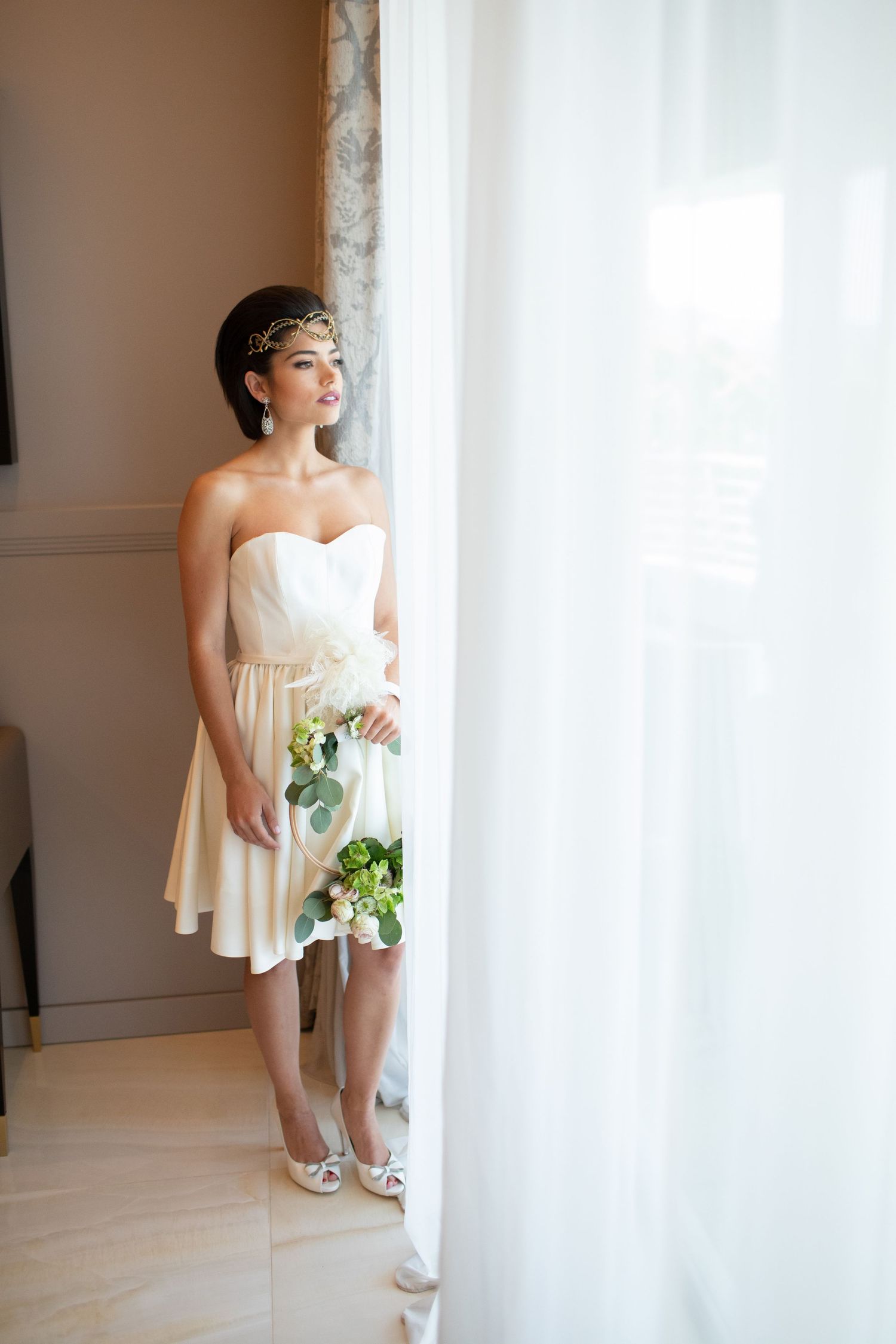 Bride in short strapless ivory dress with gold headpiece holds a bouquet while standing by sheer curtains.