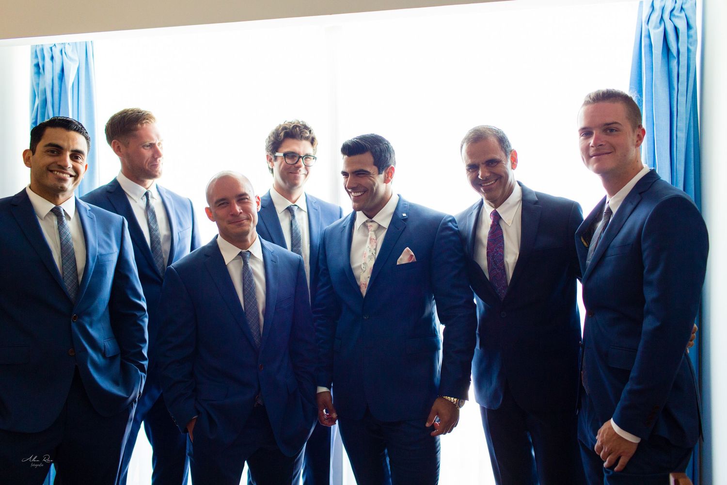 Groom surrounded by smiling groomsmen in navy blue suits posing together near bright window.