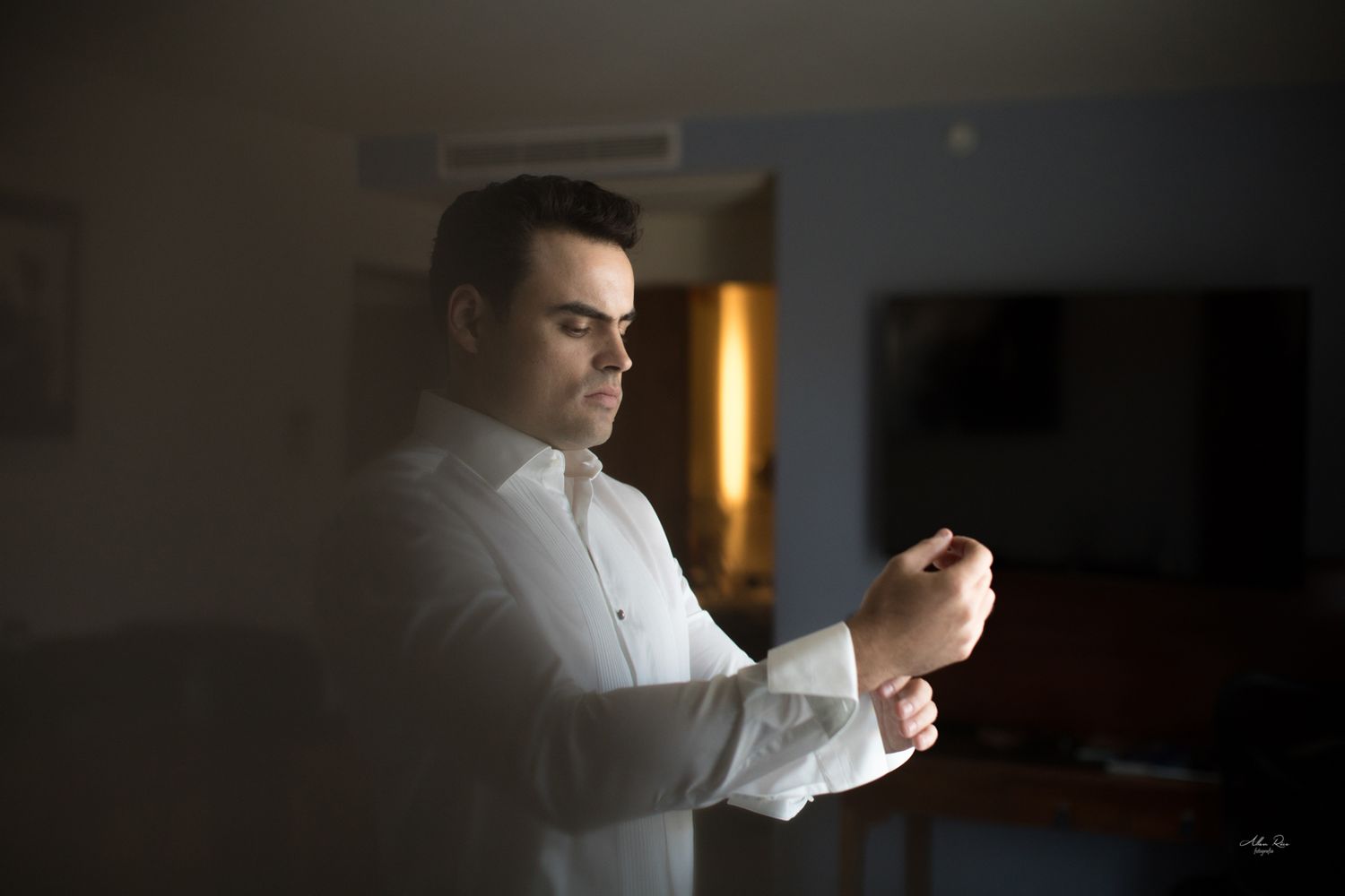 Groom in white shirt adjusting cufflinks in a dimly lit hotel room.