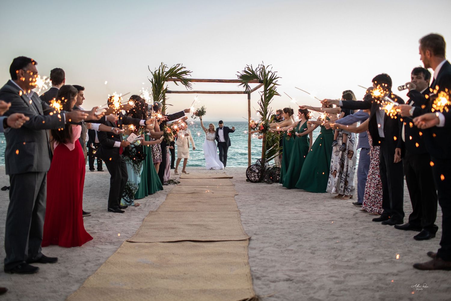 Newlyweds walk through sparkler send-off tunnel with guests in elegant attire at beach wedding.