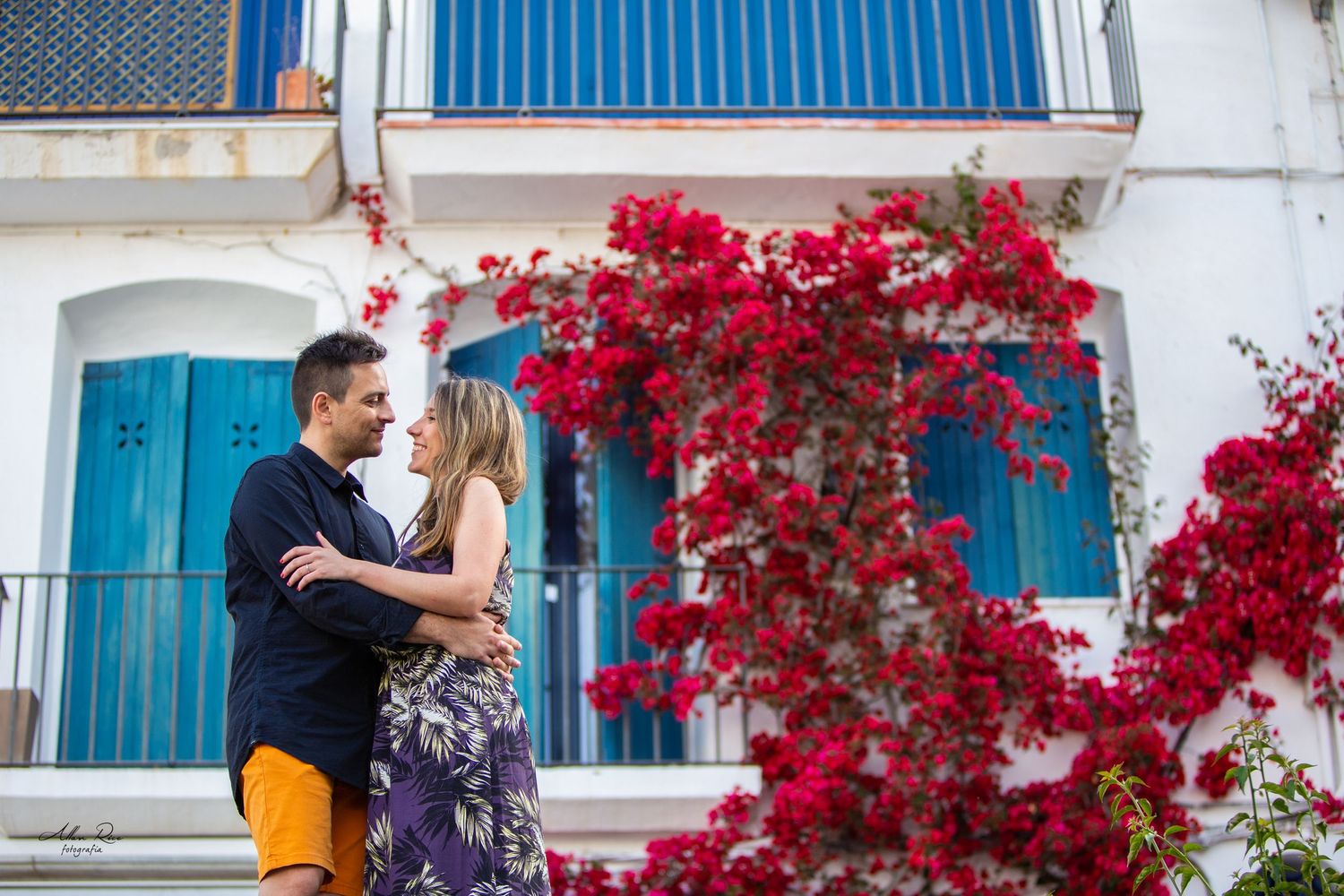 Couple embracing romantically surrounded by vibrant red bougainvillea at a white Mediterranean villa.
