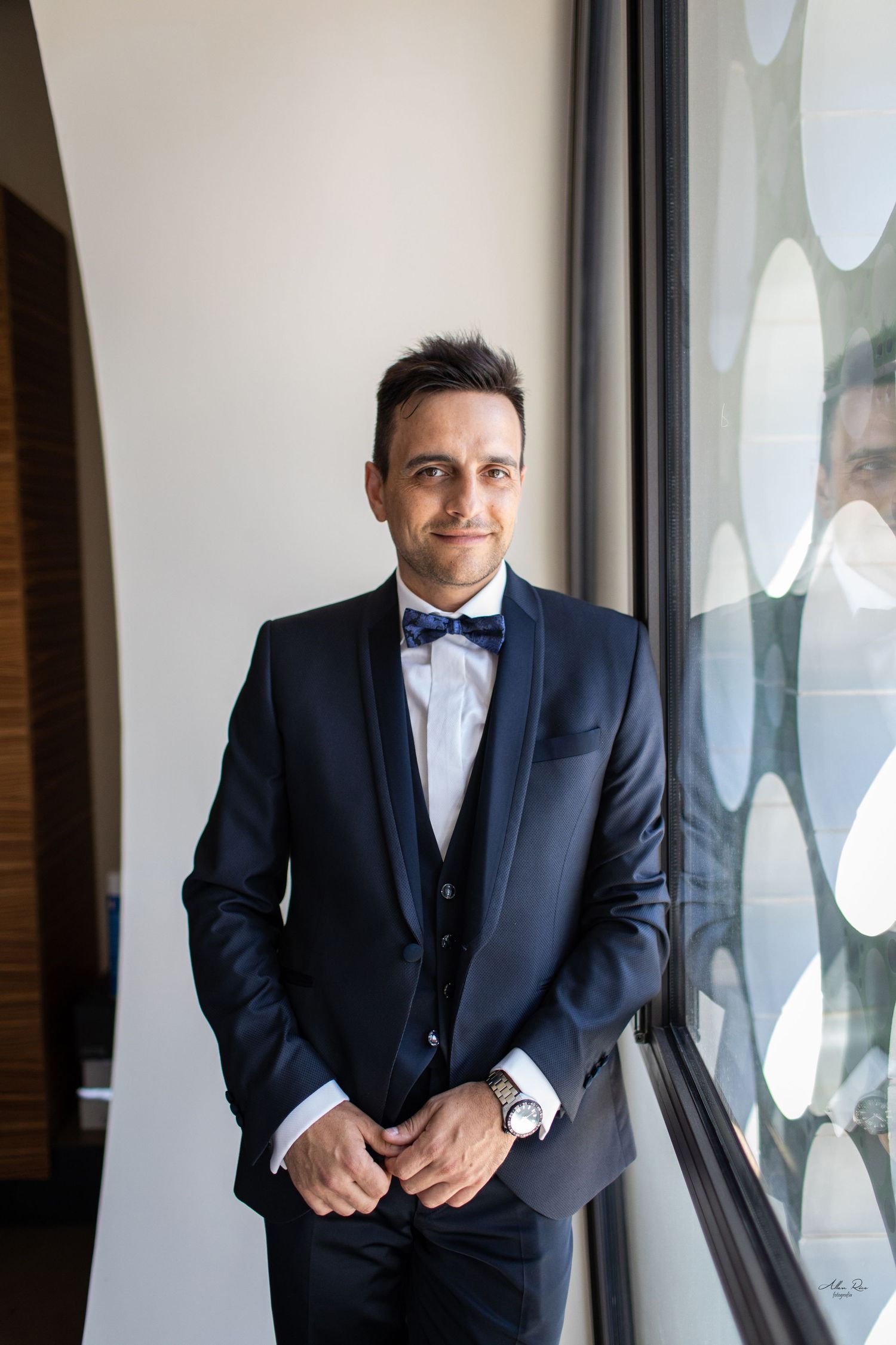 Elegant groom in navy tuxedo with bow tie standing by a window smiling.