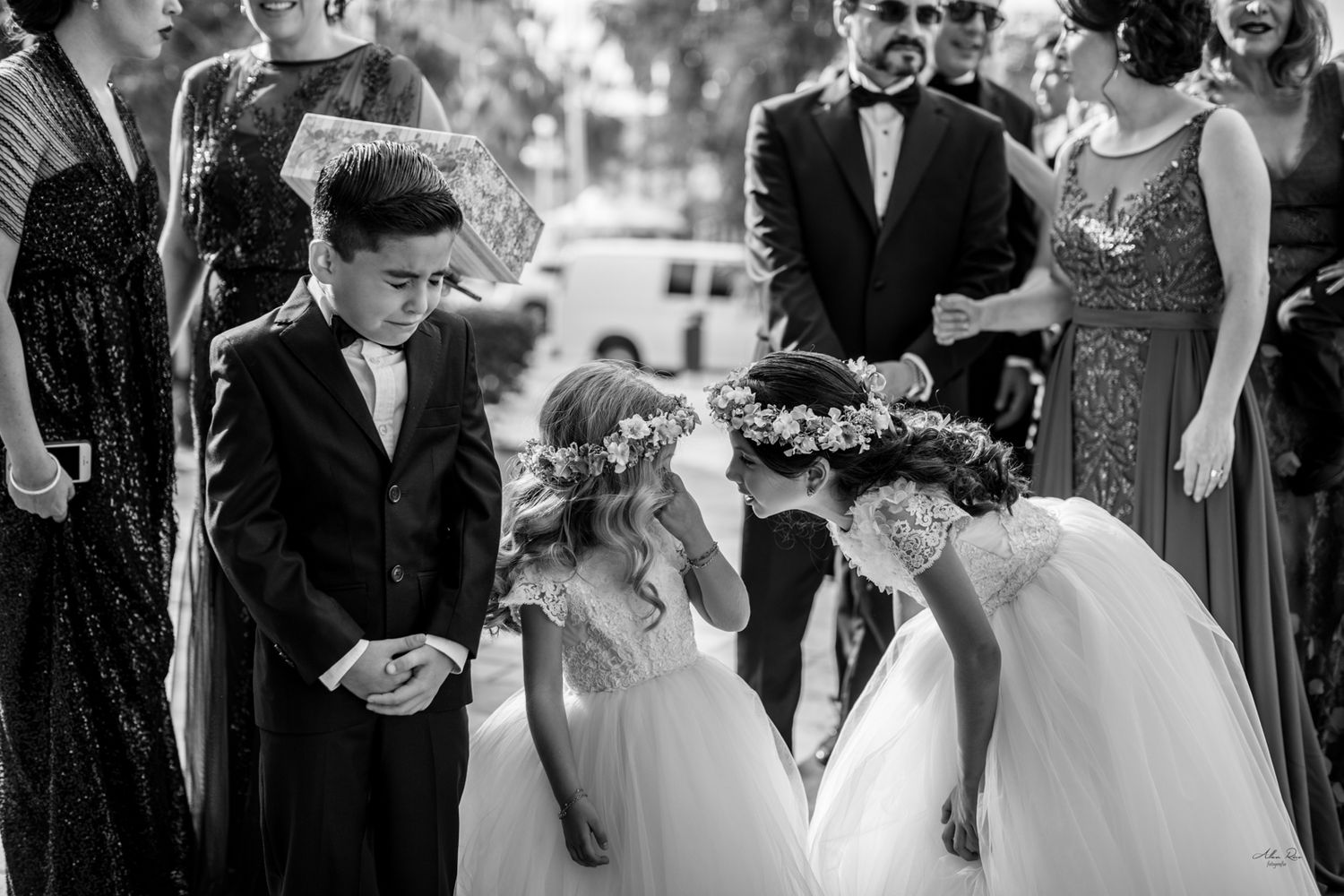 Flower girls in white dresses with floral crowns greeting ring bearer at outdoor wedding.