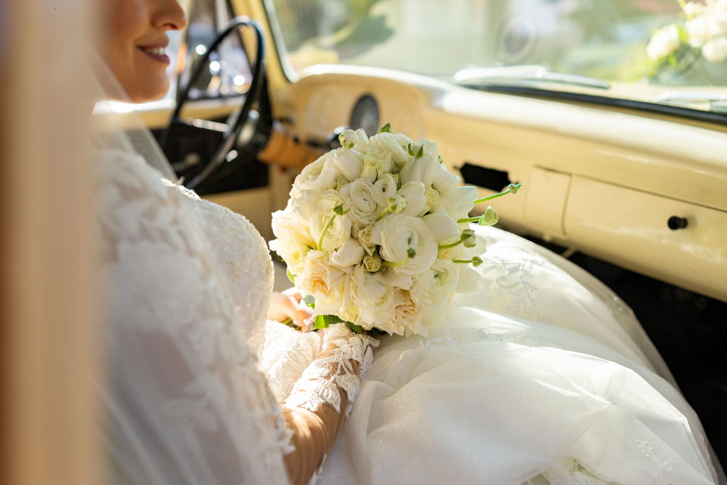 Bride in white lace gown holding elegant white flower bouquet inside a vintage yellow car.