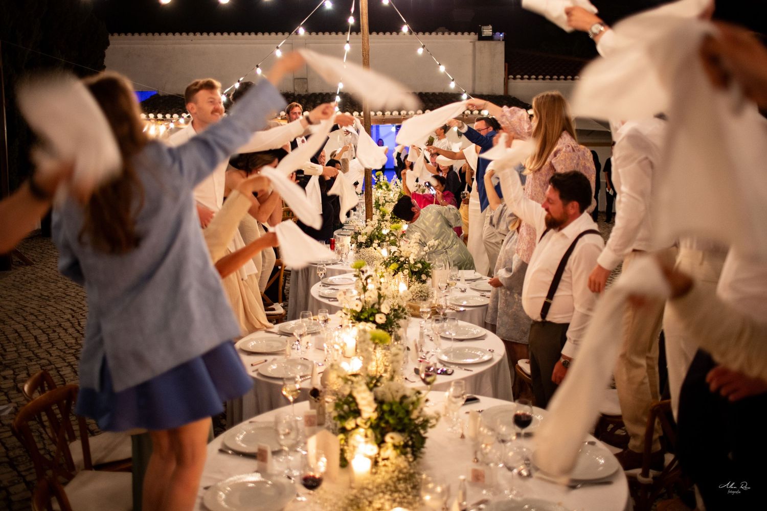 Wedding guests wave white napkins over candlelit reception dinner table at night.