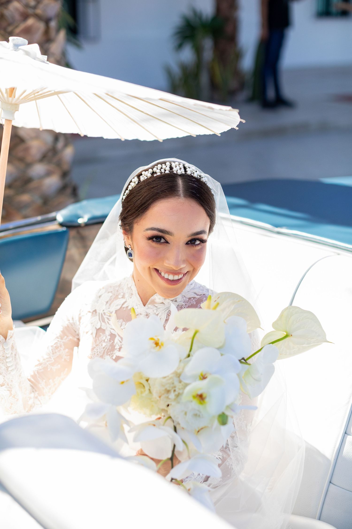 Smiling bride with white orchid bouquet under parasol in convertible car.