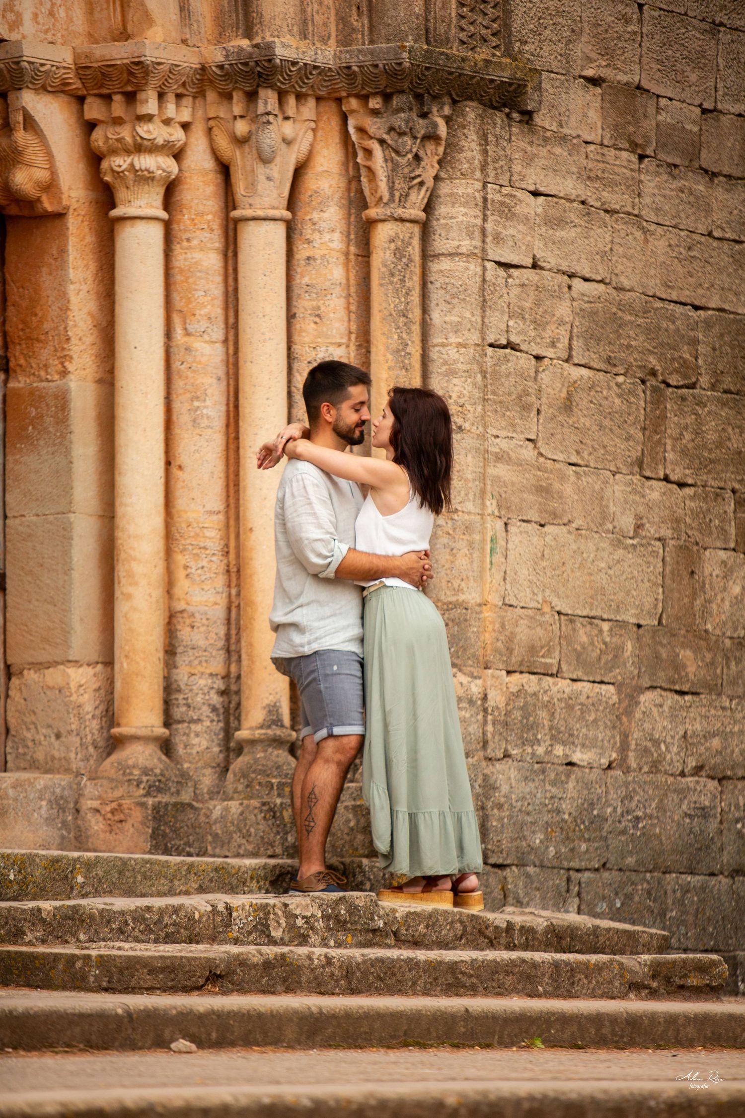 Couple embracing romantically on stone steps beside ornate medieval columns of an ancient stone church.