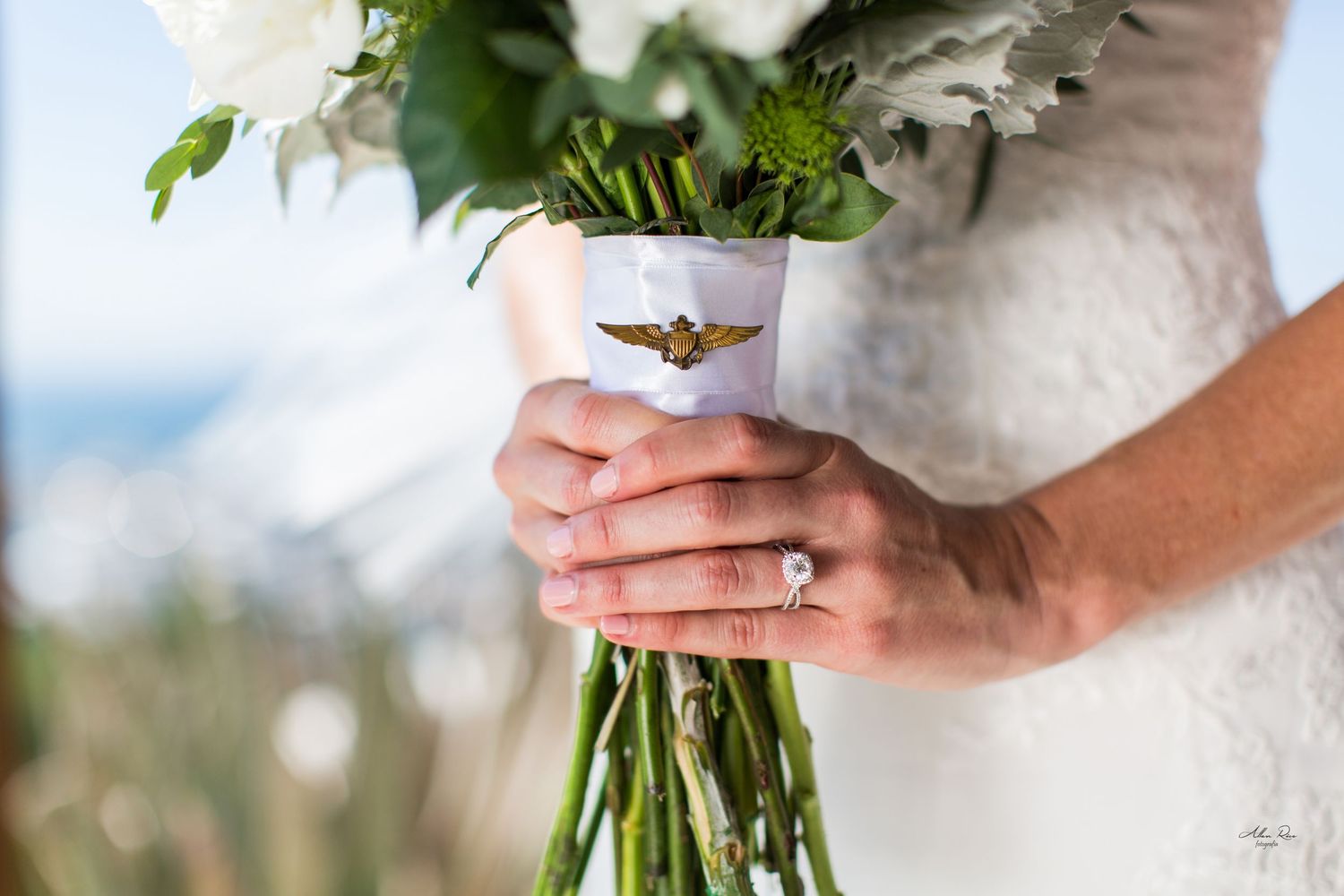 Close-up of bride holding white floral bouquet with gold aviator pin and diamond ring.