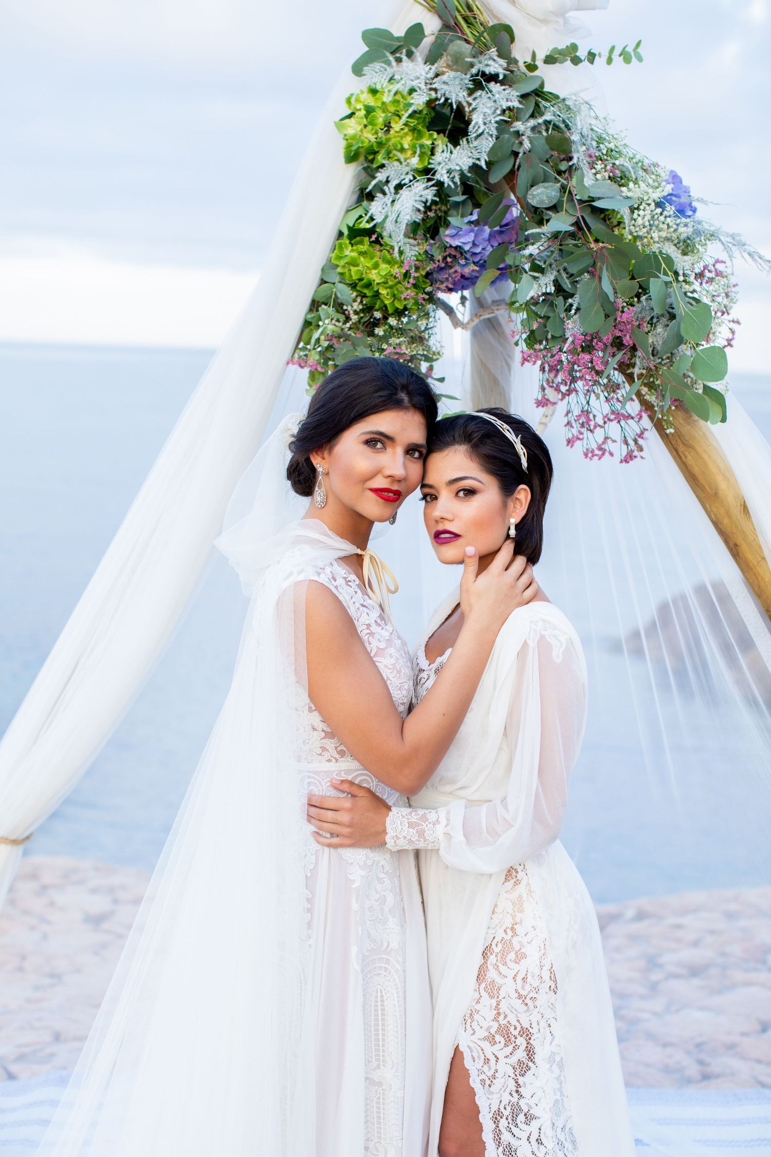Two brides in white lace gowns embracing under a floral arch decorated with purple and green blooms.