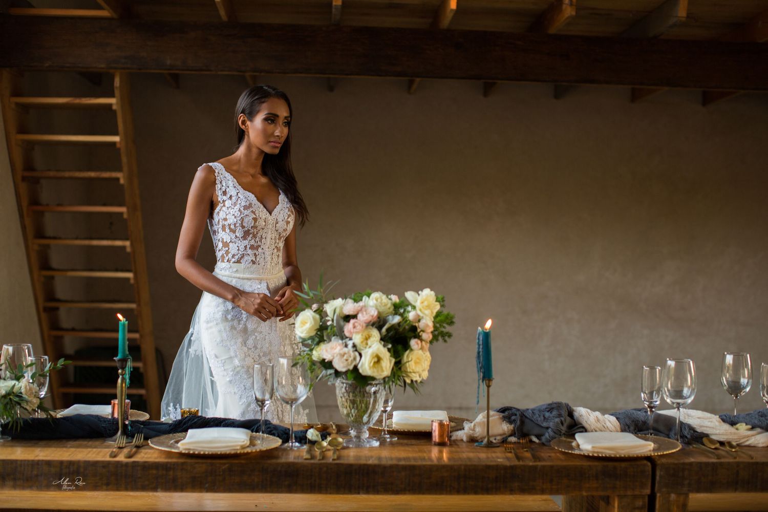 Elegant bride in lace gown standing at decorated reception table with floral centerpiece indoors.
