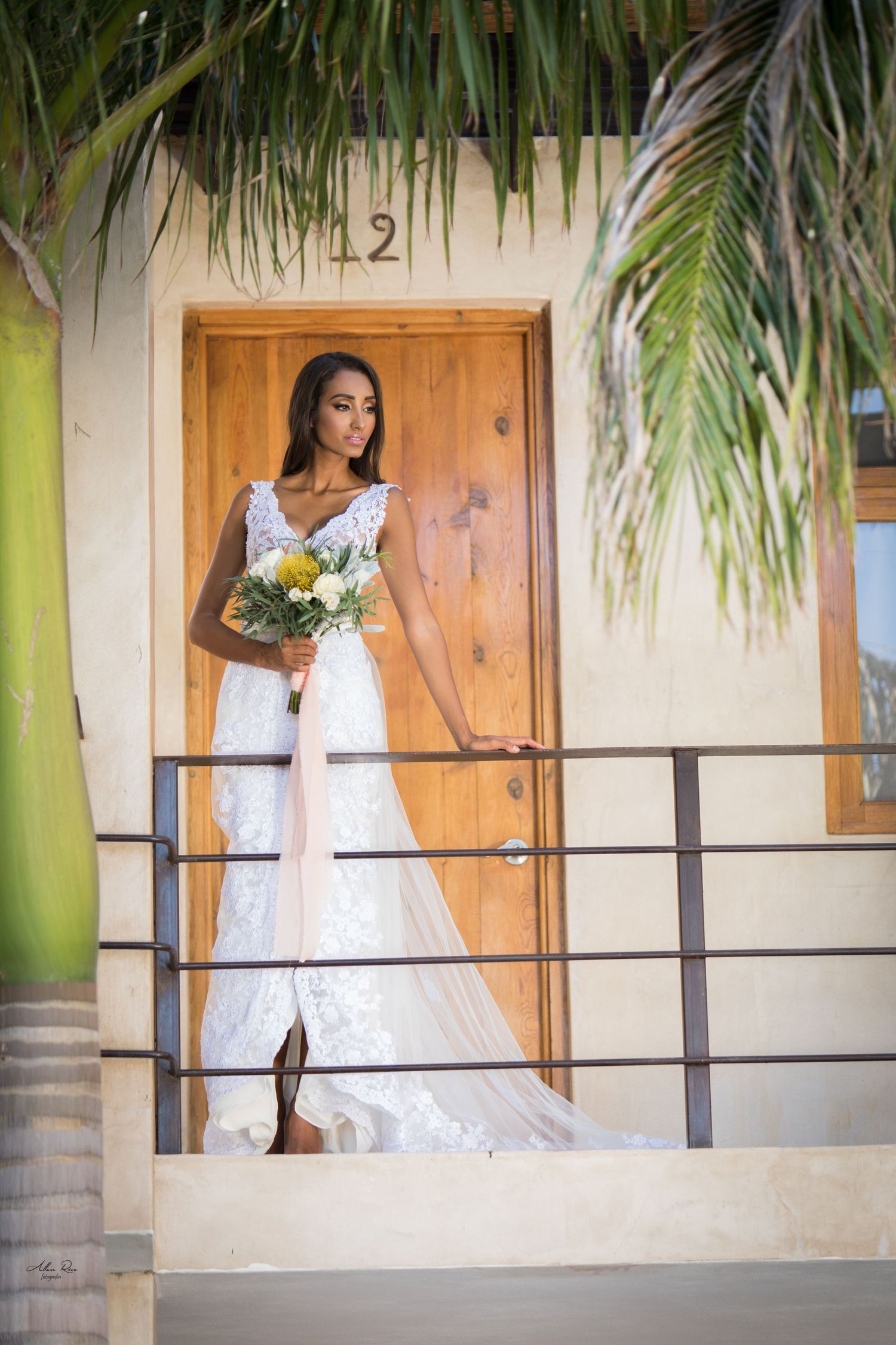 Bride in lace wedding dress holding bouquet stands by wooden door numbered 12 on sunny balcony.