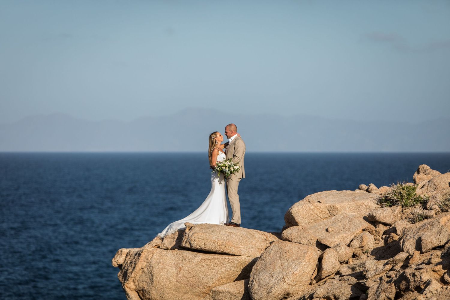 Bride and groom embrace on rugged rocky cliffs with deep blue ocean stretching behind them.