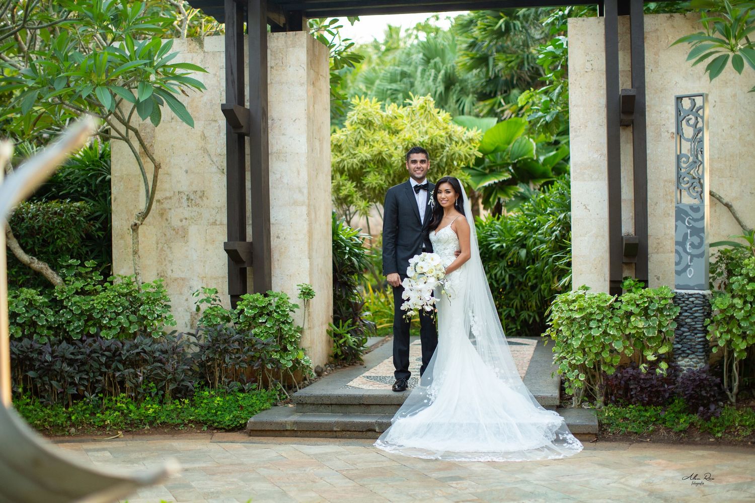 Bride and groom pose elegantly beneath stone pillars surrounded by lush tropical greenery.