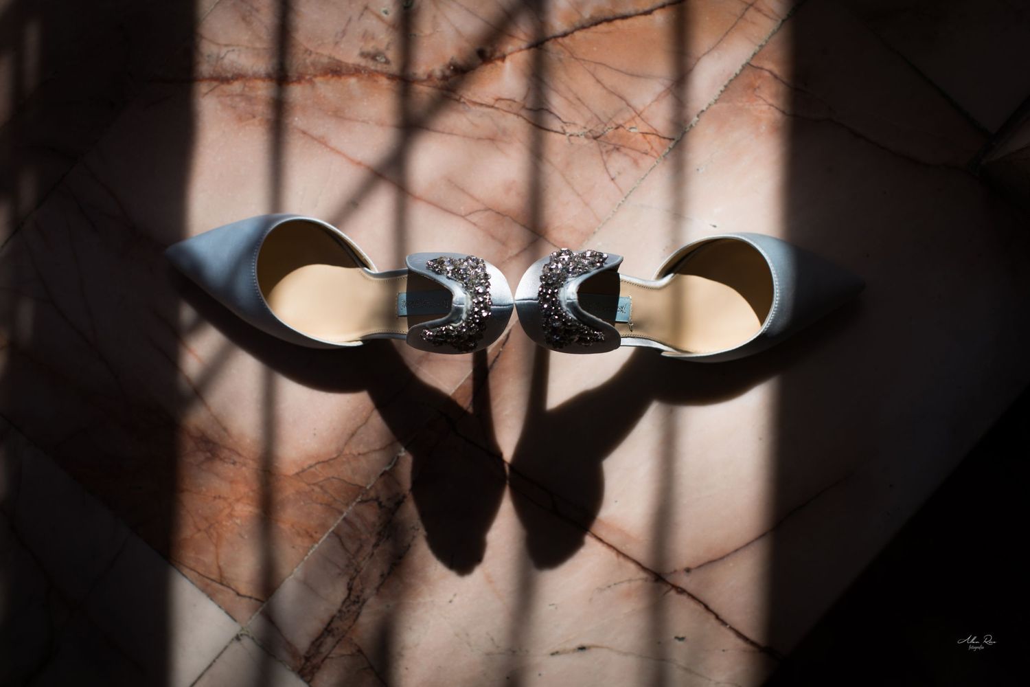 Elegant silver bridal heels with rhinestone bow detail casting dramatic shadows on marble surface.