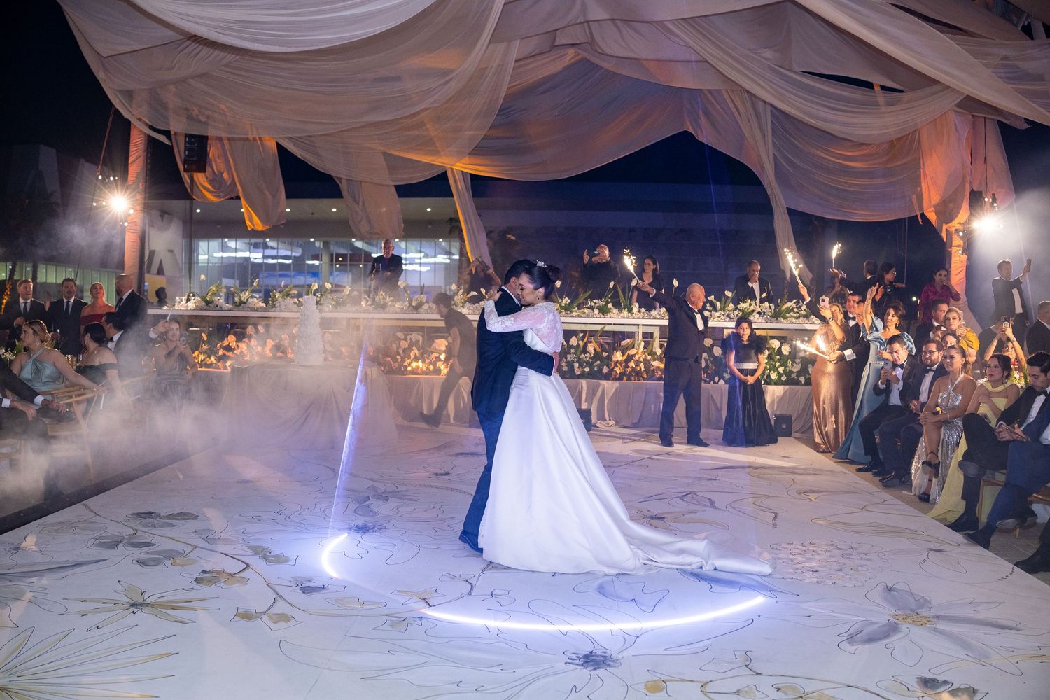 Couple shares first dance on illuminated circular floor under draped canopy with sparkler-holding guests.