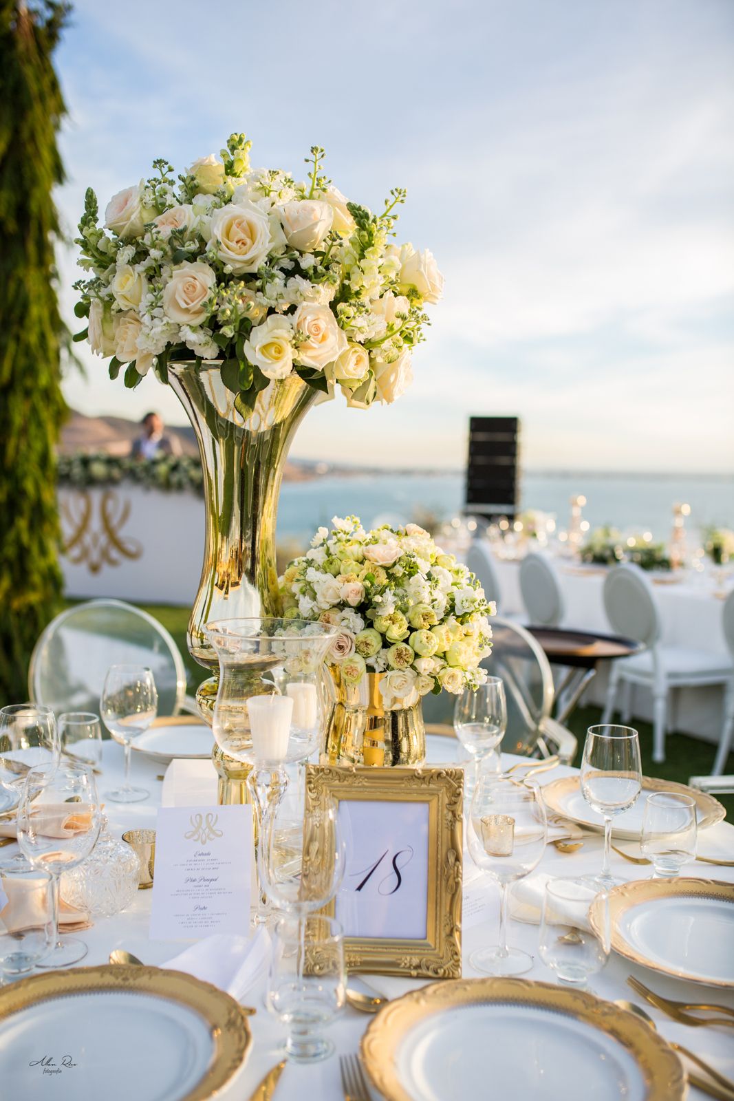 Luxurious outdoor wedding reception table with tall white rose centerpieces, gold-rimmed plates, and ocean backdrop.