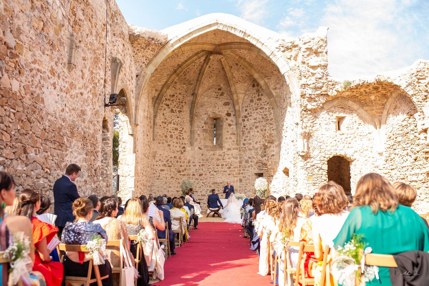 Wedding ceremony held in a stunning ancient stone ruins with guests seated along a red carpet aisle.