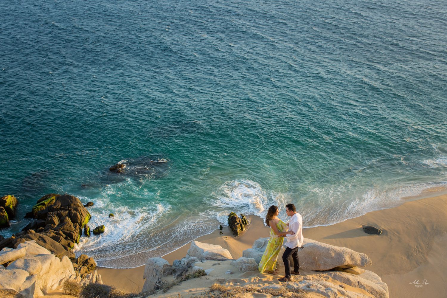 Couple standing on rocky cliffs above turquoise ocean waves during a romantic engagement shoot.