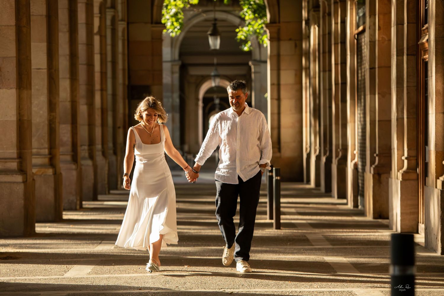 Couple walking hand in hand through a sunlit classical stone archway colonnade.