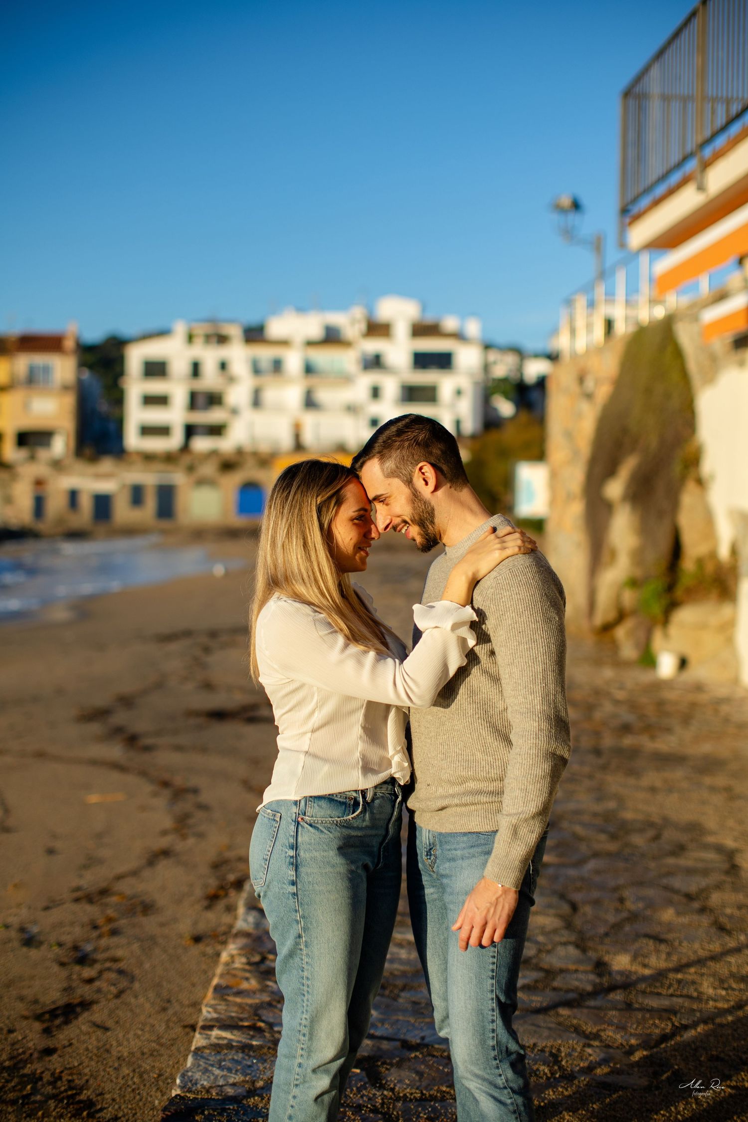 Happy couple embracing on a sunny beach with whitewashed buildings and rocky coastline behind them.
