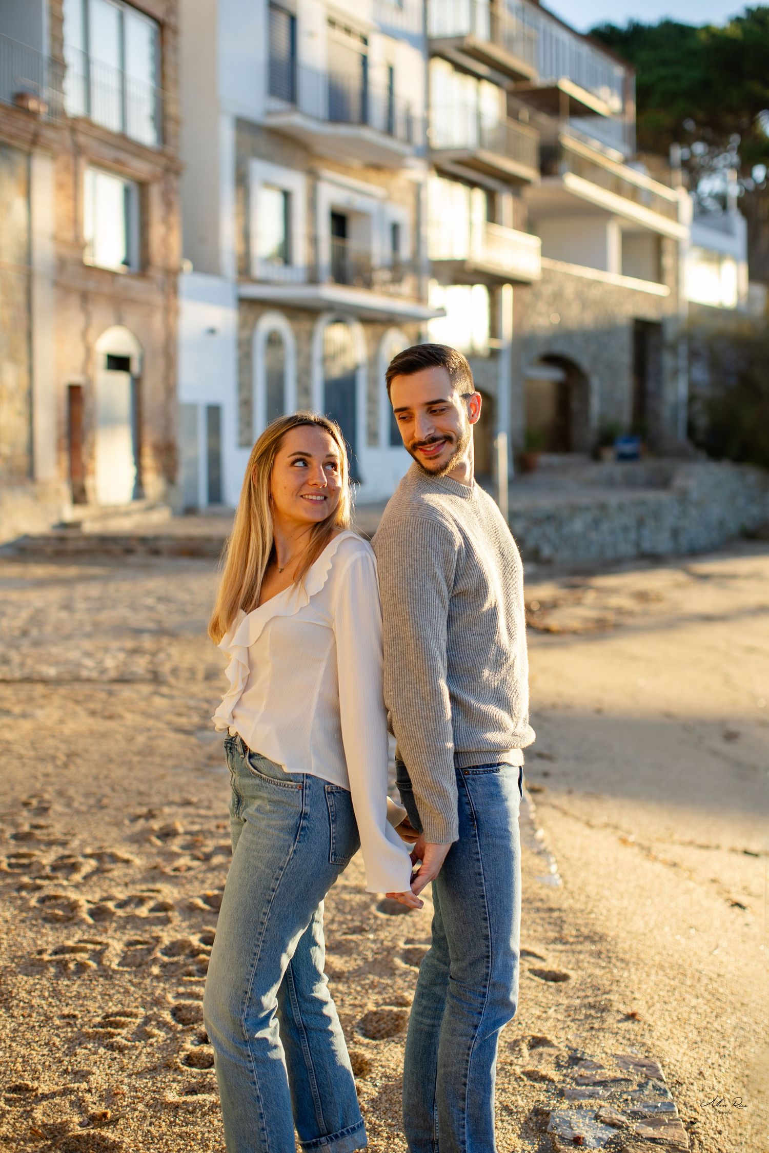 Smiling couple holding hands and posing on a sandy beach near whitewashed buildings.