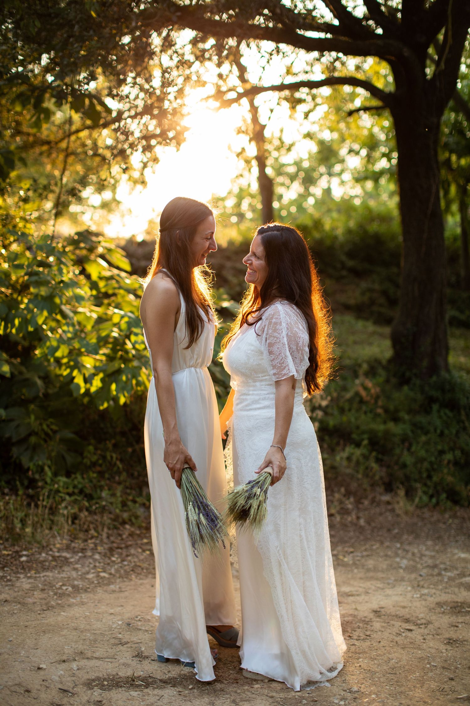 Two brides in white gowns hold wildflower bouquets and smile at each other in golden forest light.