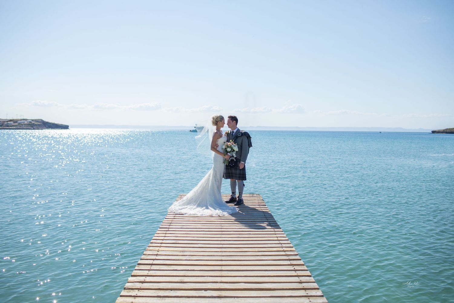 Bride and groom kiss on a wooden pier overlooking stunning turquoise ocean waters.