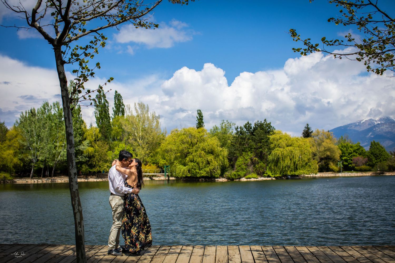 Couple sharing romantic kiss on wooden dock by lake with mountains and blue sky backdrop.