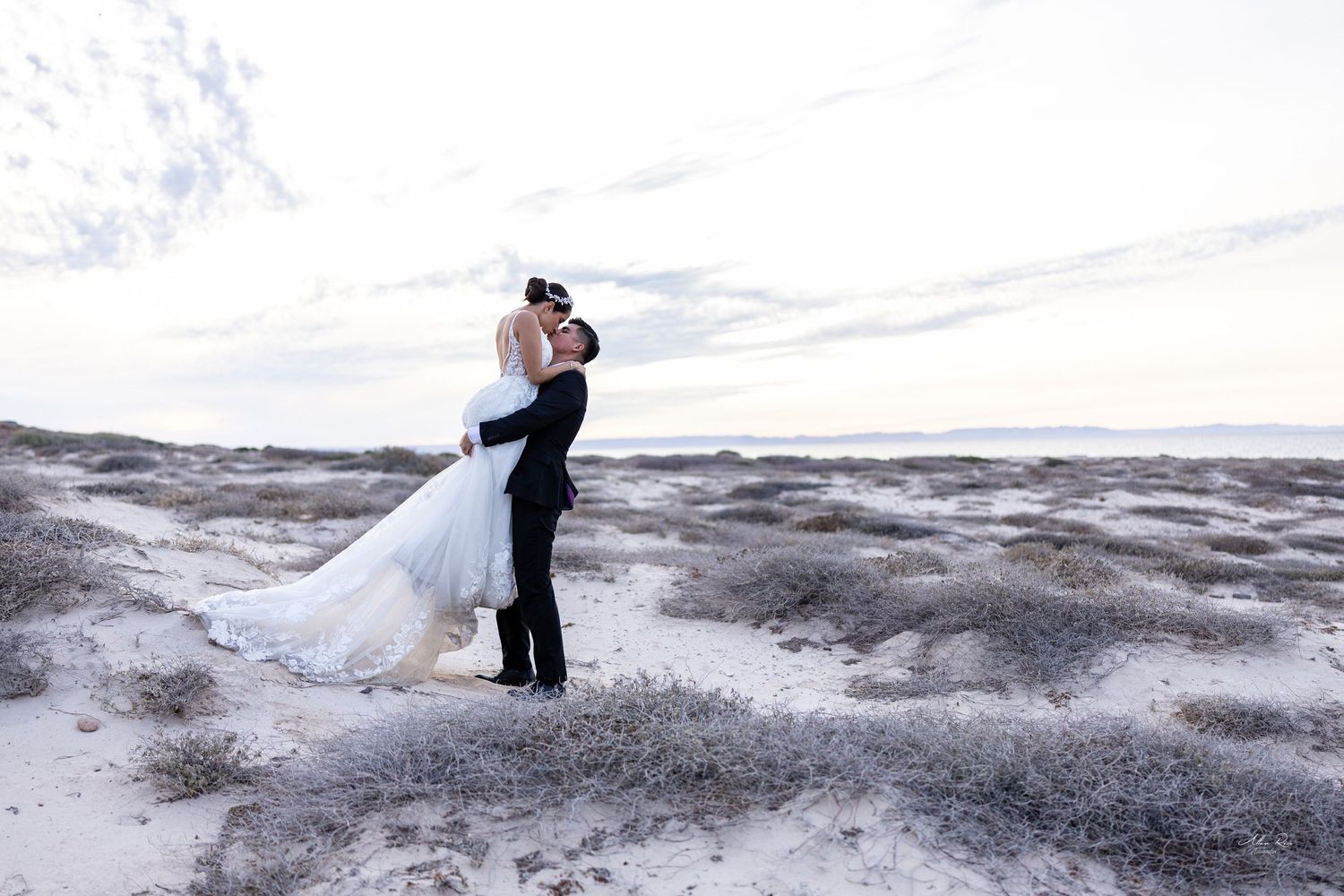 Groom lifting bride in white gown on sandy dunes under a pastel sky.