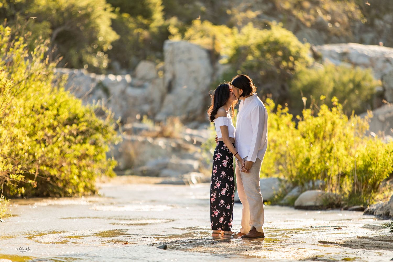 Couple shares a tender moment kissing at golden hour in a scenic outdoor rocky landscape with wildflowers.