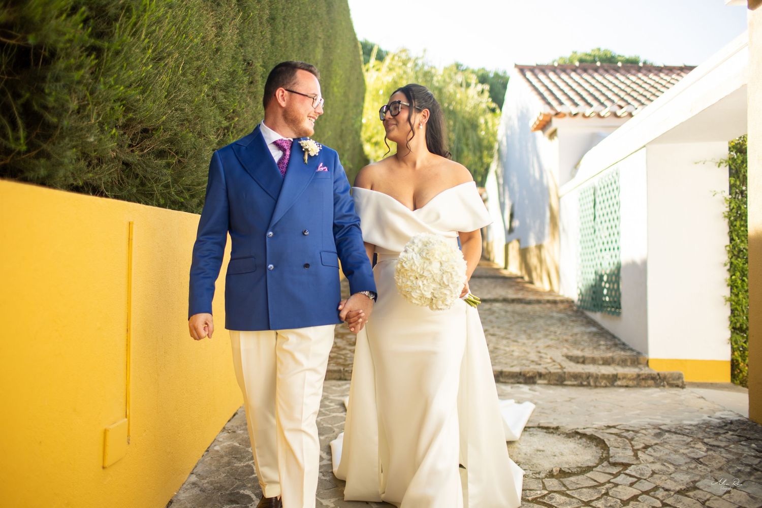 Bride in white off-shoulder gown and groom in blue suit walk hand in hand on a sunny path.