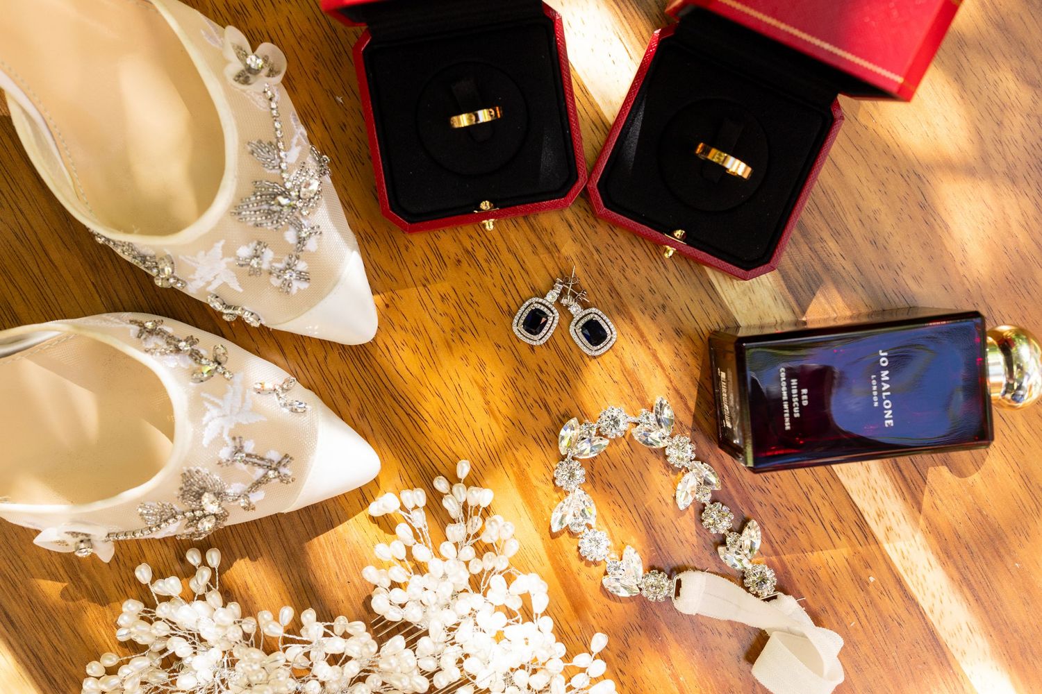 Flat lay of wedding accessories including rings, heels, earrings, bracelet, and cologne on wood.