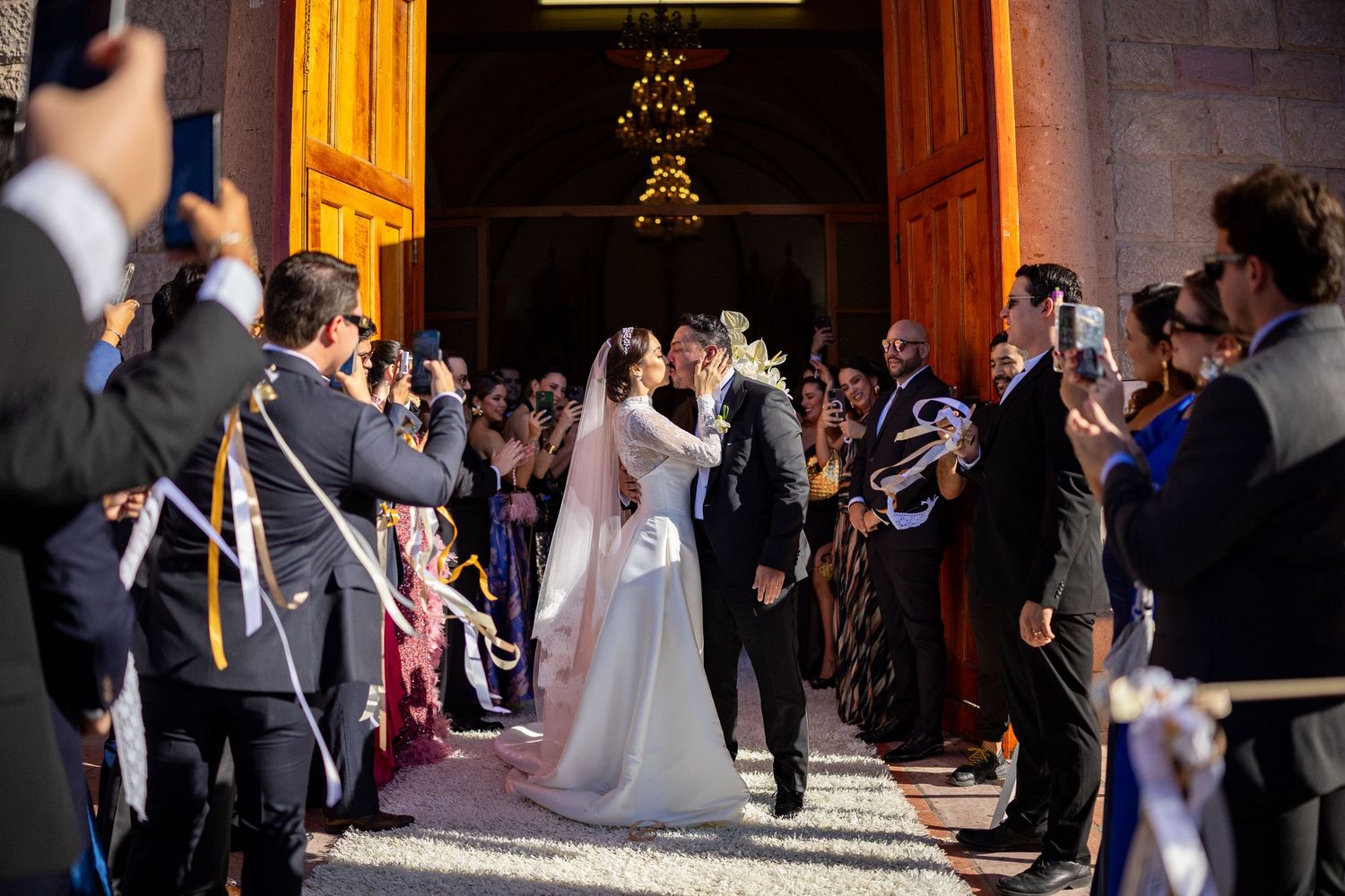 Newlyweds sharing a kiss while exiting a grand church amid cheering wedding guests.
