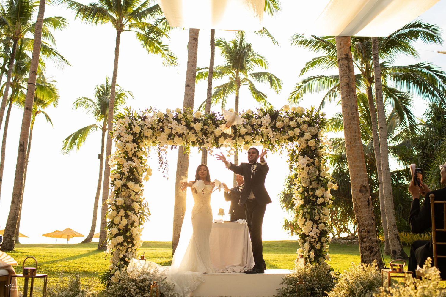 Bride and groom celebrating under a lush white floral wedding arch with palm trees and golden sunset backdrop.