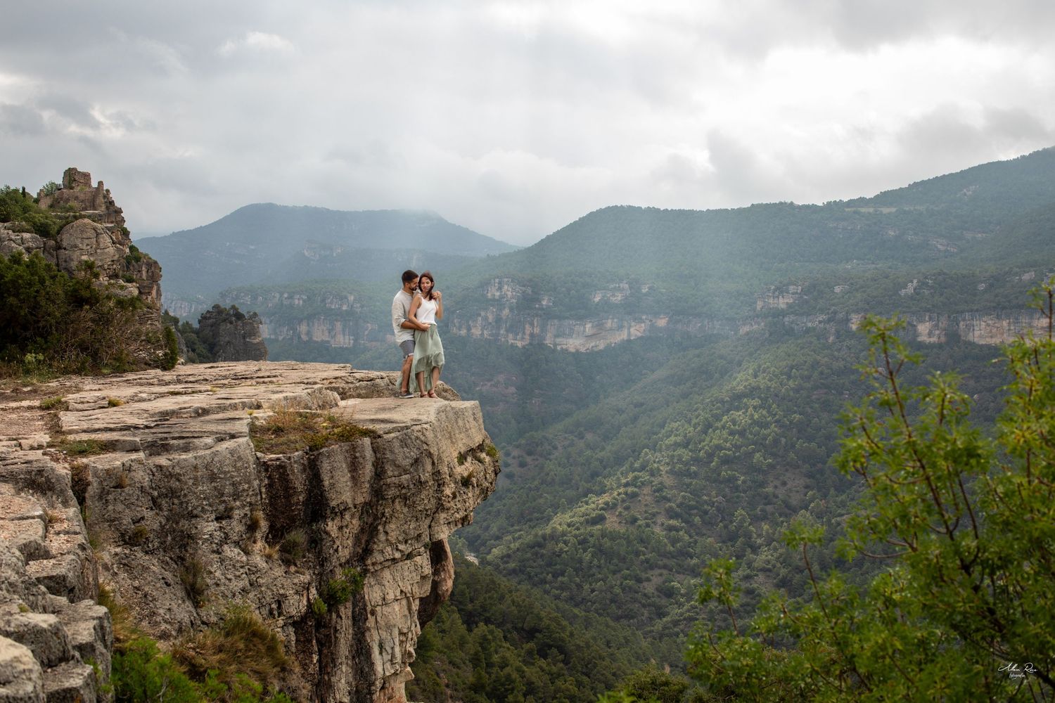 Couple kissing on a dramatic rocky cliff edge surrounded by lush green canyon and overcast mountain scenery.