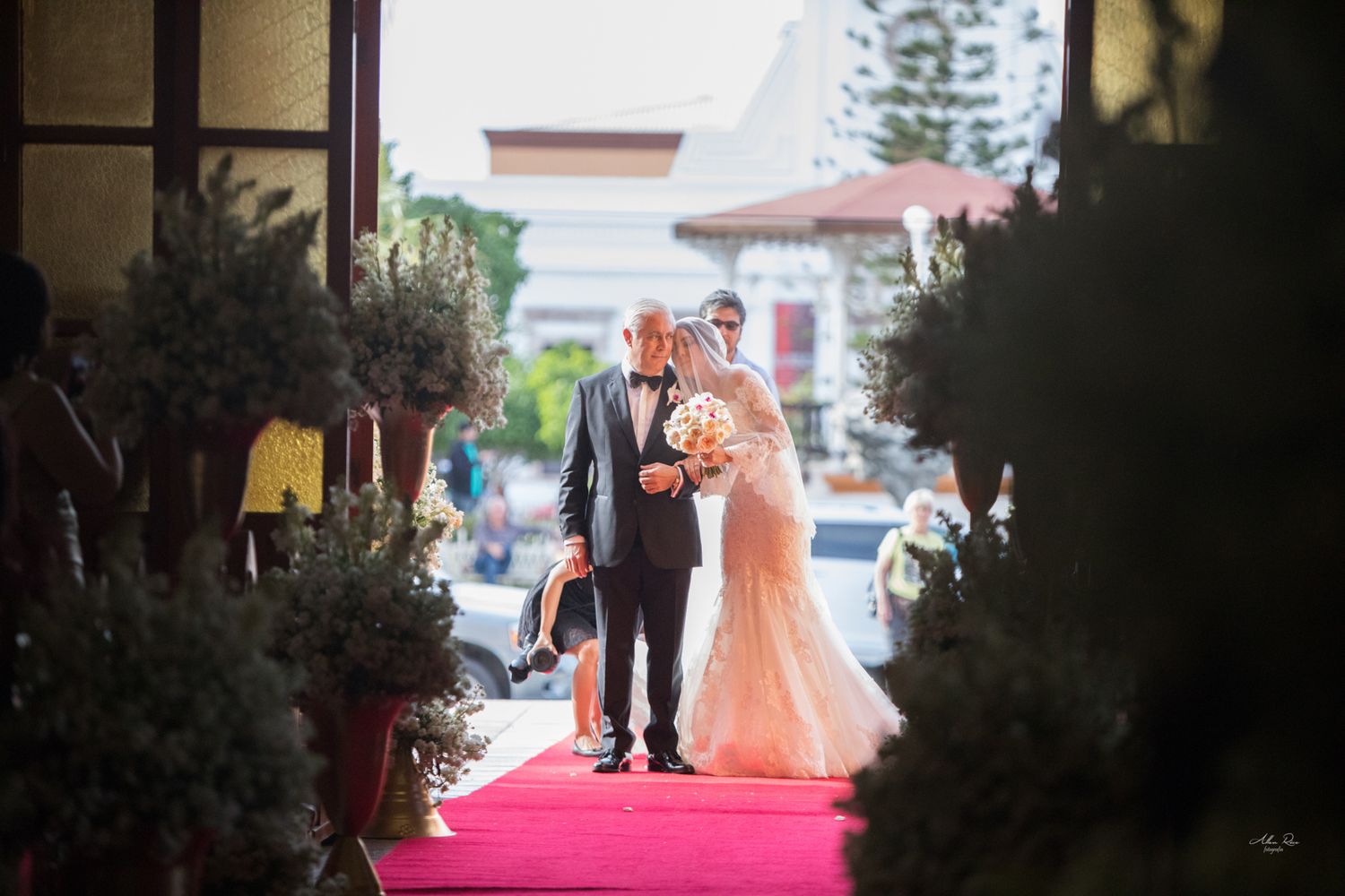 Father escorting bride in lace gown down red carpet aisle at church wedding entrance.