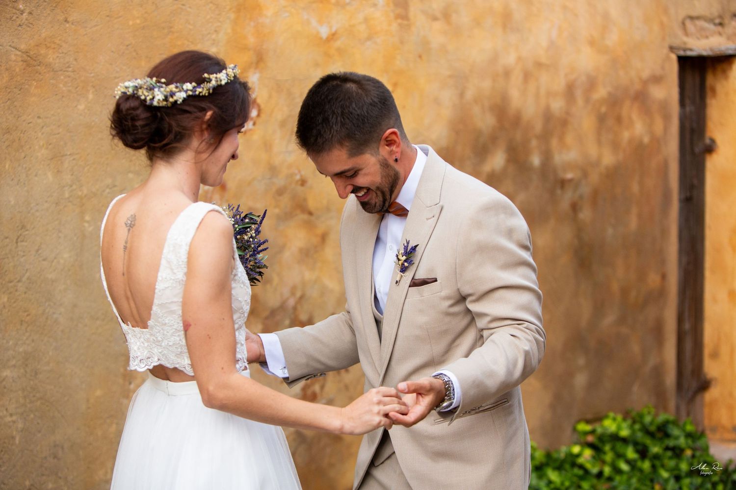 Bride and groom share ring exchange moment against warm golden weathered wall outdoors.