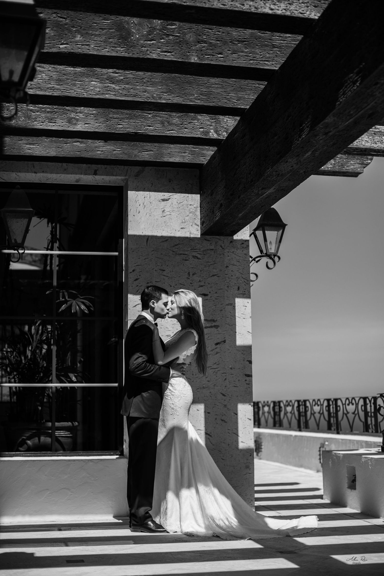 Black and white photo of bride and groom sharing a kiss on a sunlit terrace with ornate stone columns.