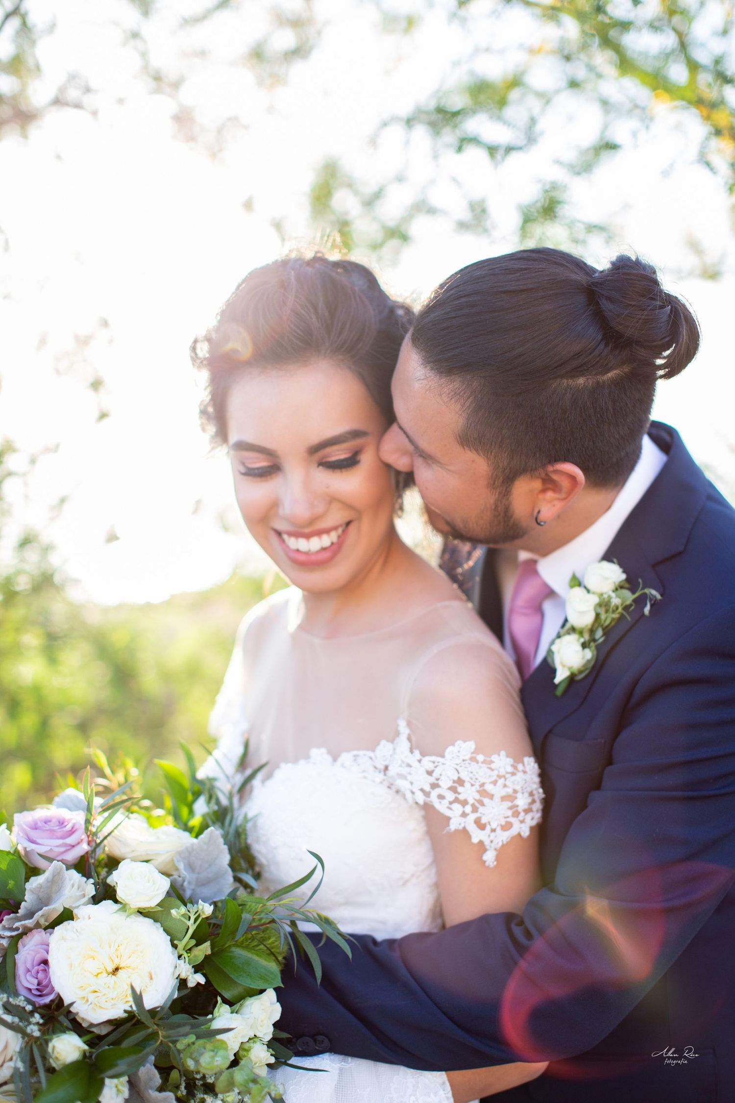 Groom tenderly kisses bride on the cheek as she smiles holding a pastel bouquet in golden sunlight.