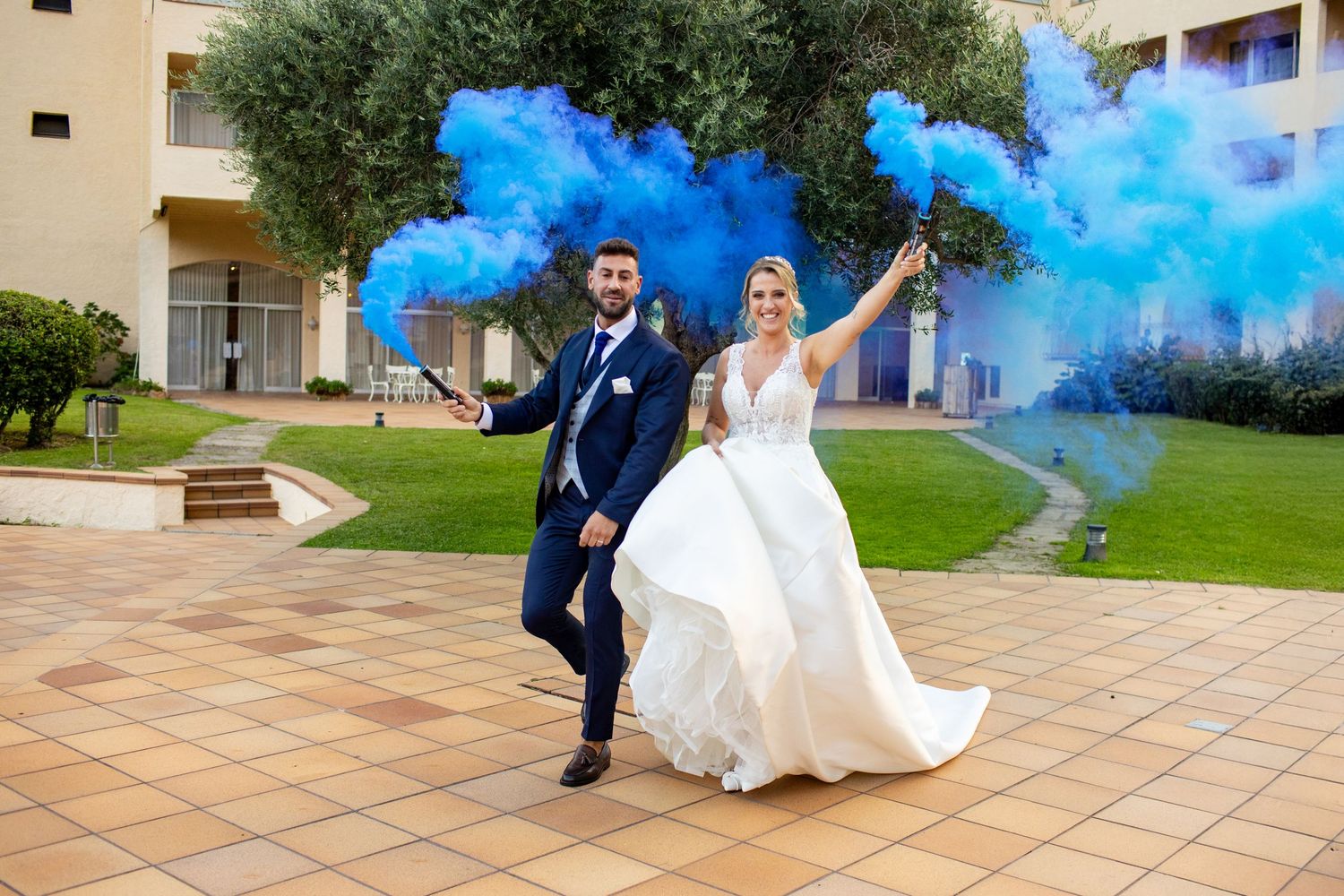 Joyful bride and groom celebrating with blue smoke bombs in outdoor wedding venue.