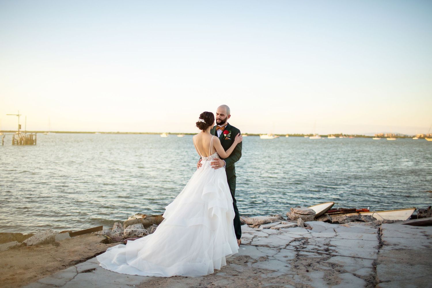 Bride and groom embracing by calm waterfront at dusk, wedding dress trailing on rocky ground.