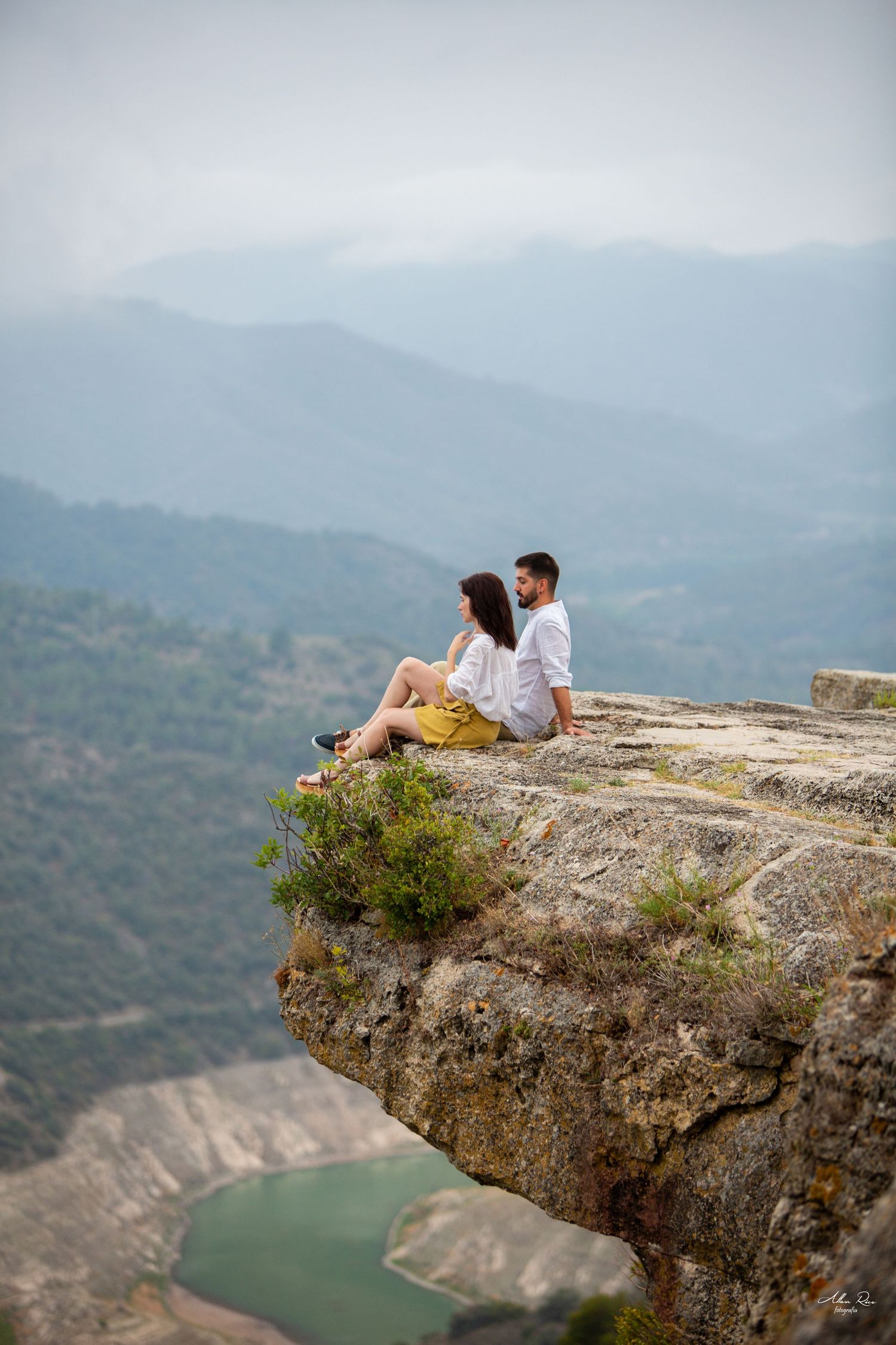 Romantic couple sitting together on a rocky cliff overlooking a turquoise river and misty mountain landscape.