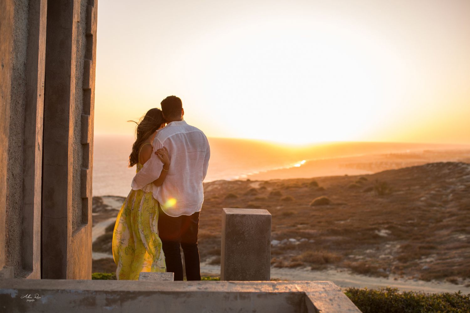 Couple sharing a romantic kiss at sunset overlooking the ocean near ancient stone columns.