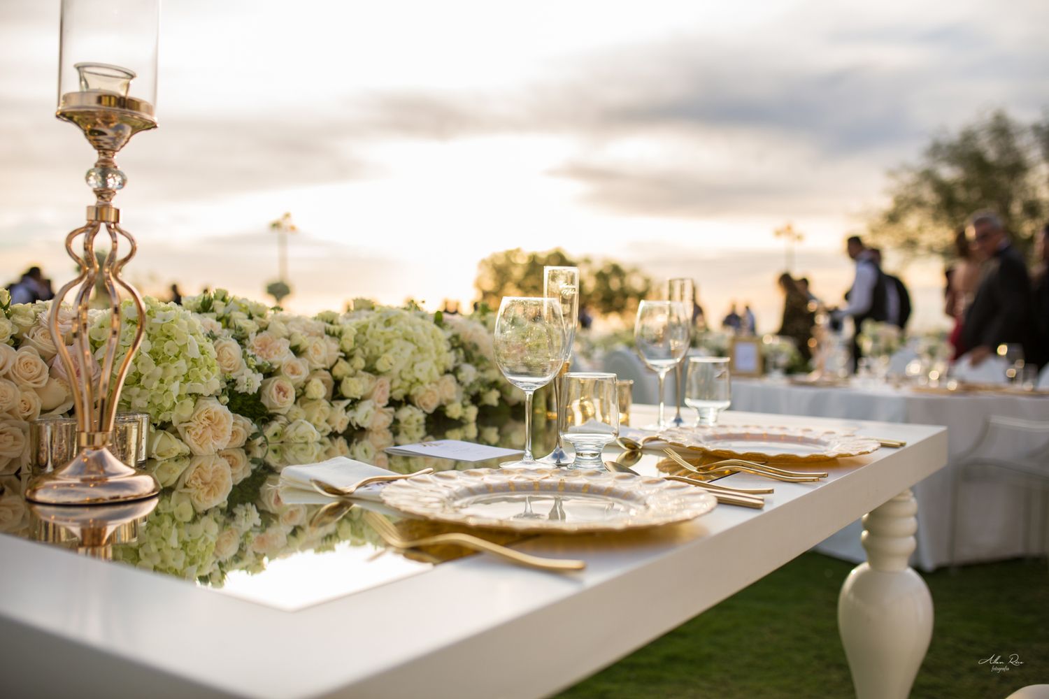 Elegant outdoor wedding reception table with gold chargers, crystal glasses, and white floral centerpieces at sunset.