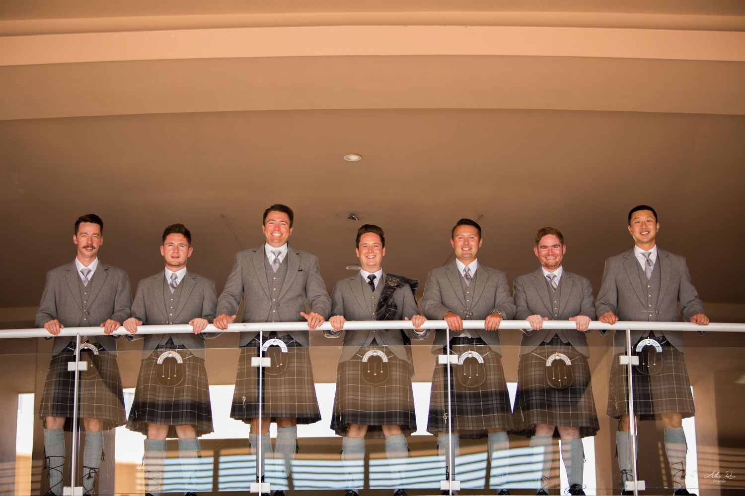 Seven groomsmen in matching gray suits and kilts posing on a balcony railing.