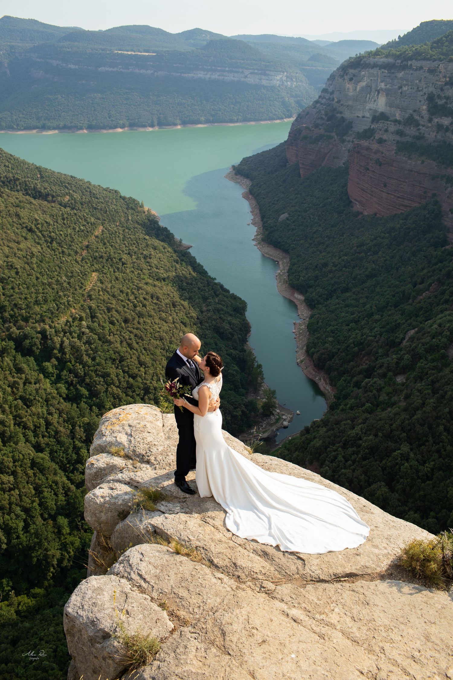 Bride and groom embracing on a rocky cliff edge with breathtaking turquoise river valley below.