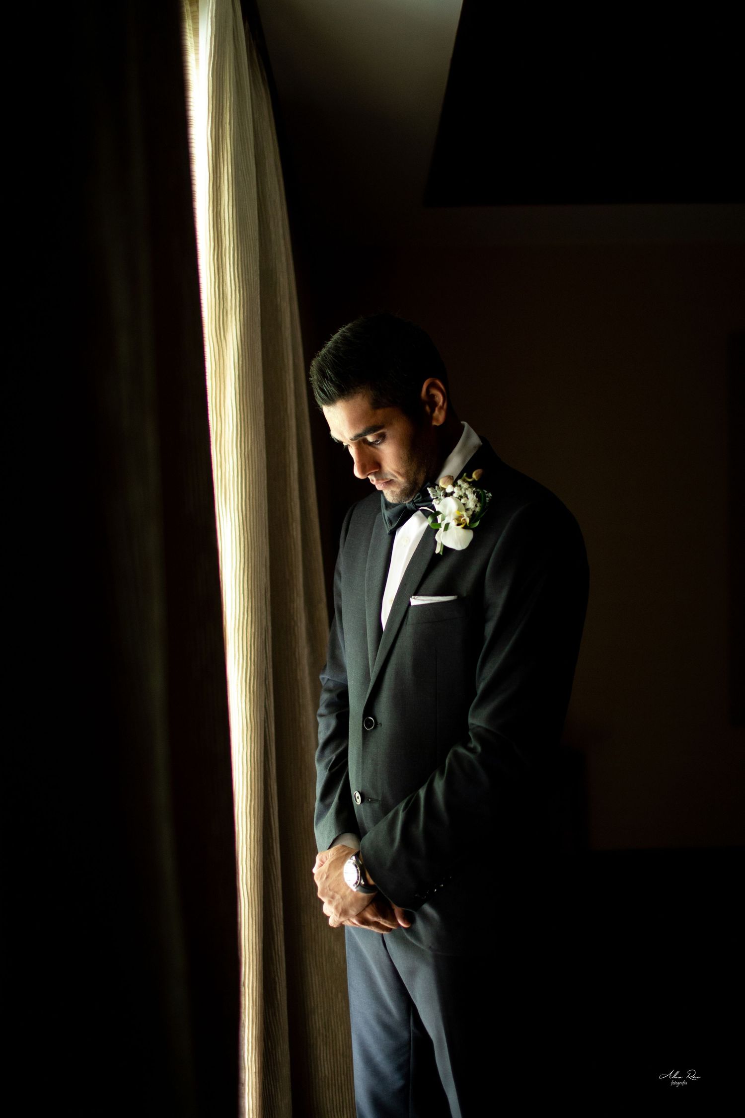Groom in black tuxedo with boutonniere stands in dramatic shadow by curtained window before ceremony.
