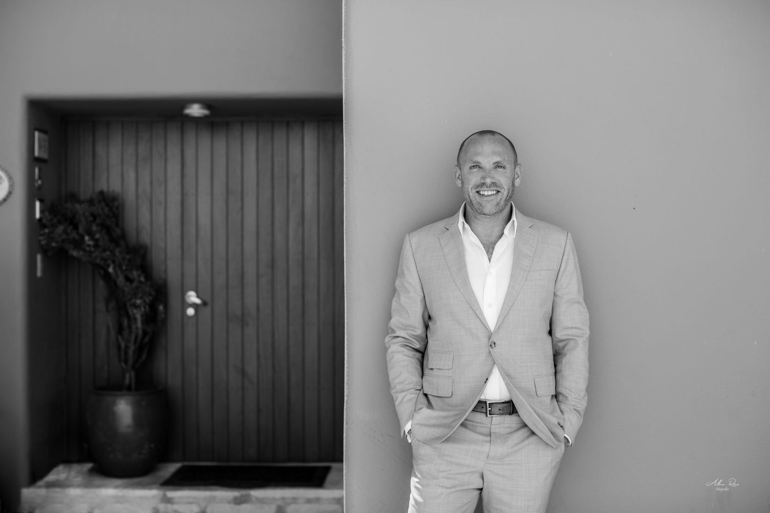 Stylish man in beige suit smiles confidently leaning against wall in black and white portrait.